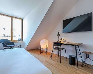 Helles Schlafzimmer mit weißer Bettwäsche, schwarzem Stuhl, Schreibtisch und großem Wandfernseher